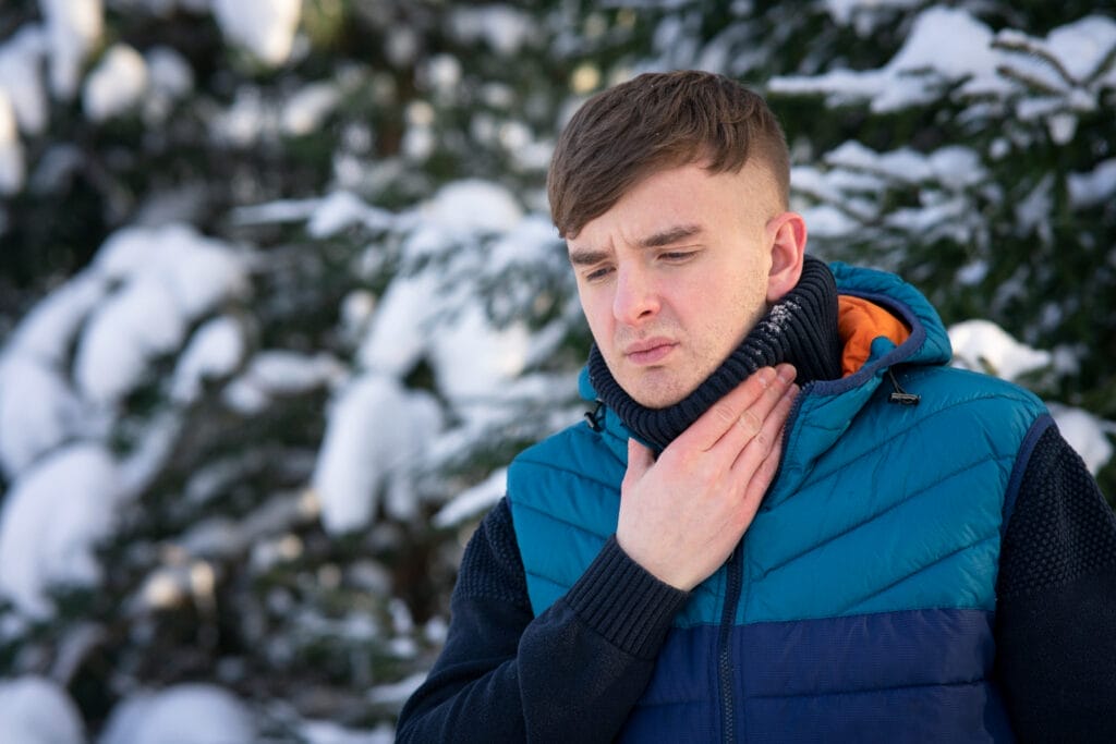 Man bundled up in a winter coat and scarf, standing outdoors in a snowy setting, visibly uncomfortable while touching his throat, suggesting a sore throat during winter.