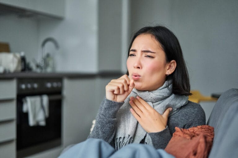 Asian woman indoors wearing a cozy scarf, holding her throat with a pained expression, suggesting discomfort from a sore throat.