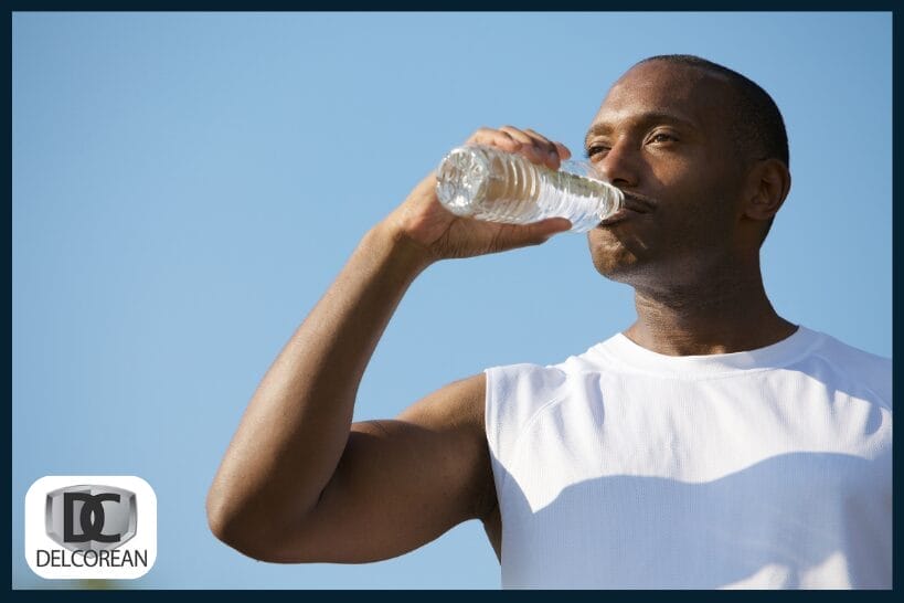 Man in a sleeveless white shirt drinks from a water bottle outdoors against a clear blue sky, highlighting hydration and electrolyte support.